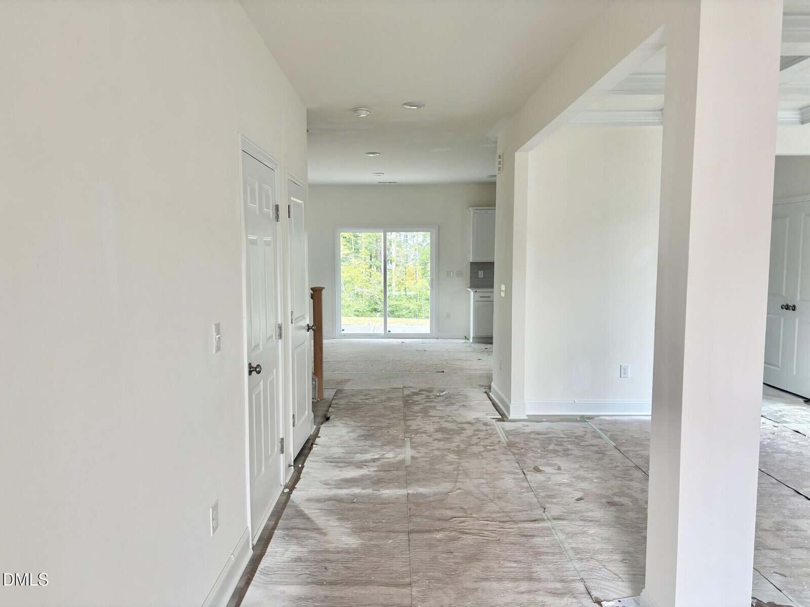 A hallway in a new, unfurnished house with white walls and doors, leading to a room with large glass doors that open to a green outdoor area. The floor appears unfinished or covered for protection.