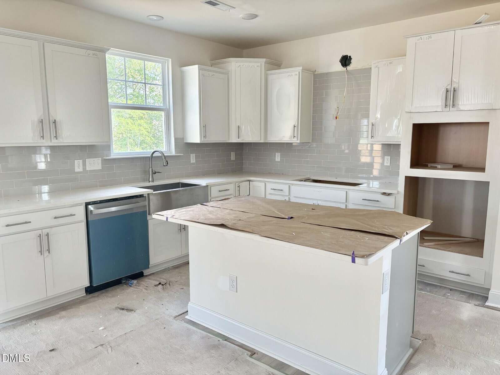 A modern kitchen under construction with white cabinets, light gray subway tile backsplash, an island covered with cardboard, and a window above the sink. Some appliances are missing or not yet installed.