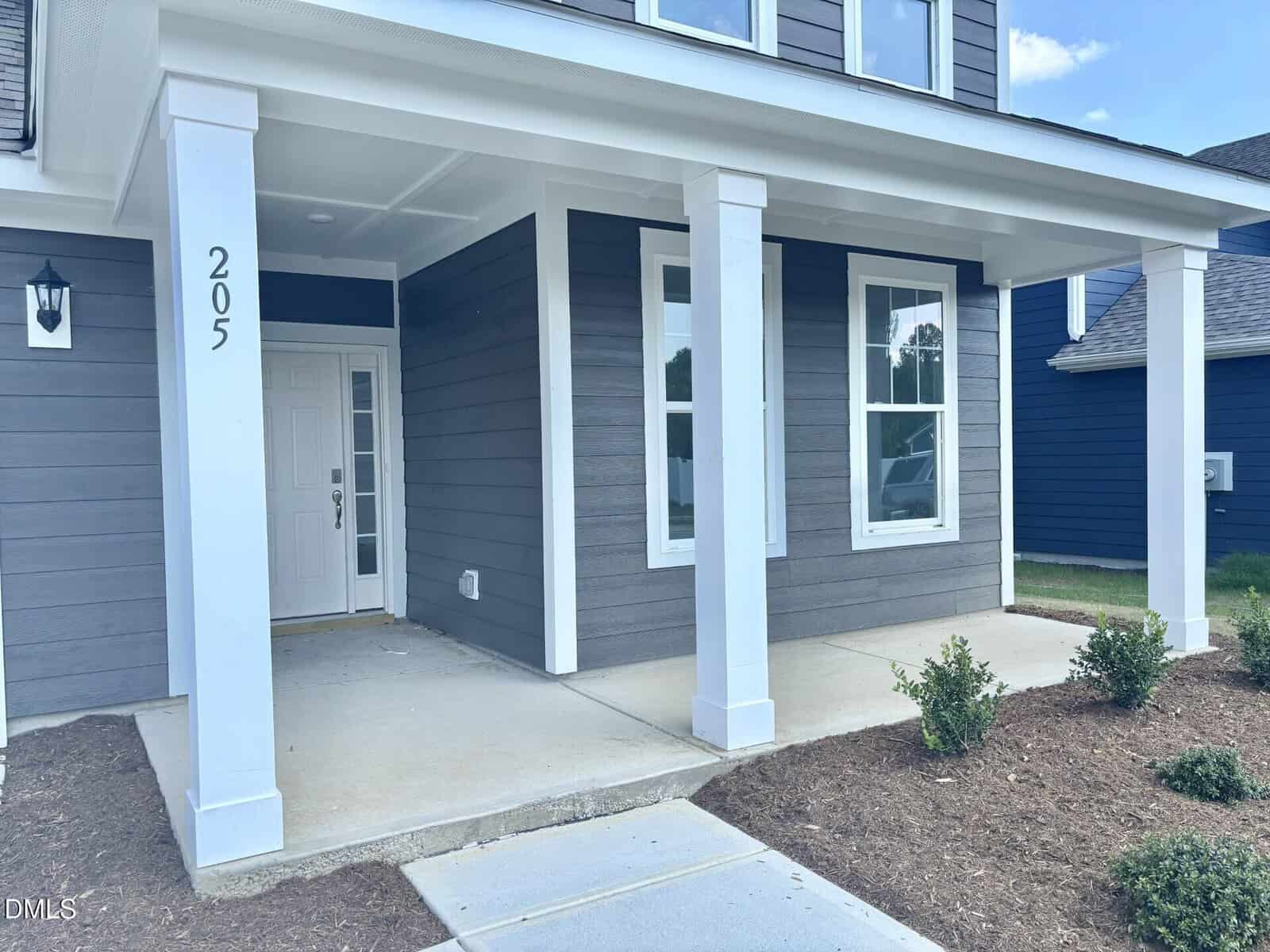 A gray house with white trim features two white columns supporting a covered front porch. The house number 205 is visible on a column. A white front door and several windows are present. Some small shrubs border the porch.