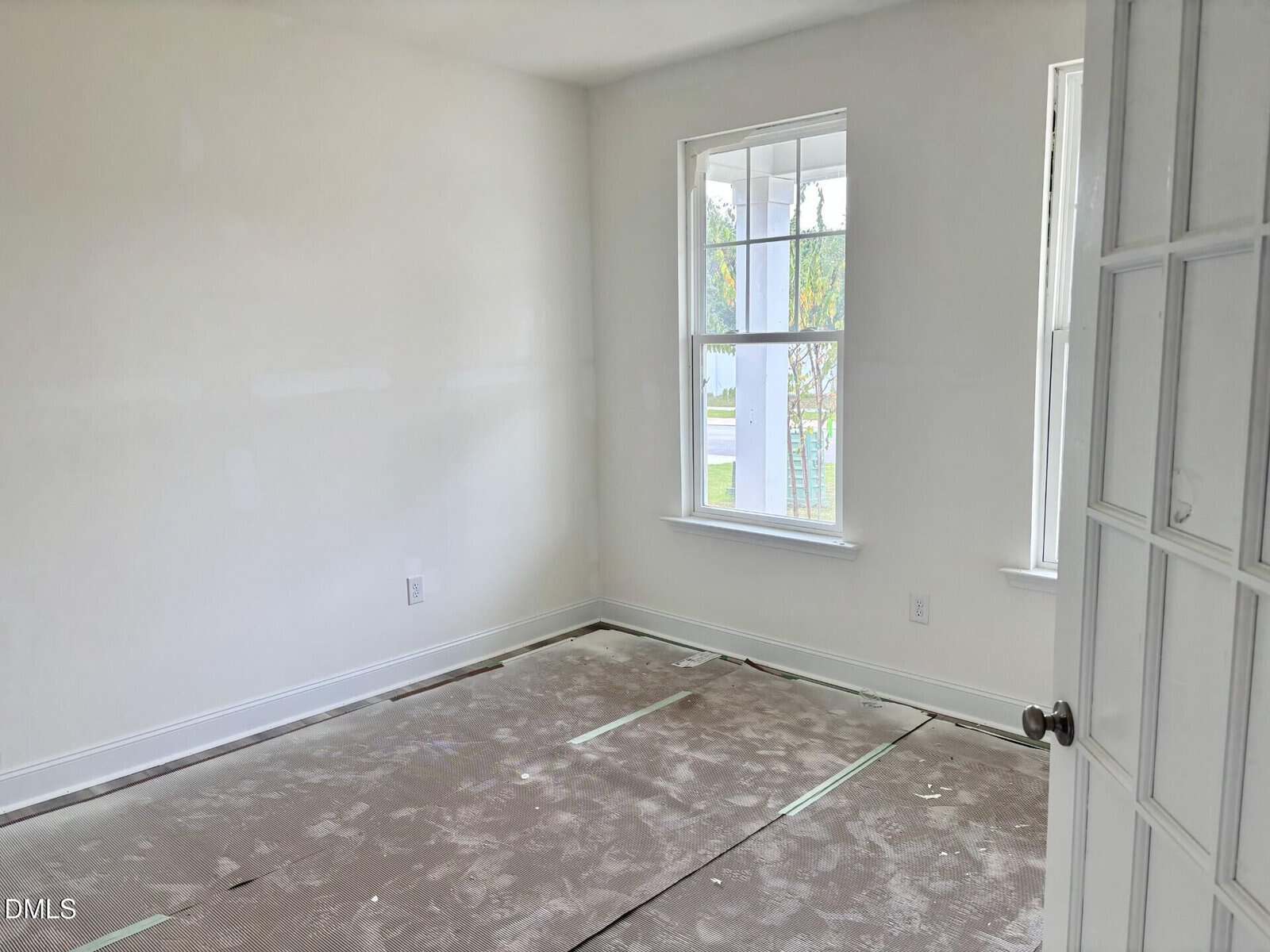 An empty room with white walls and two large windows letting in natural light. The floor is unfinished and covered with protective material. A door with glass panels is partially open.