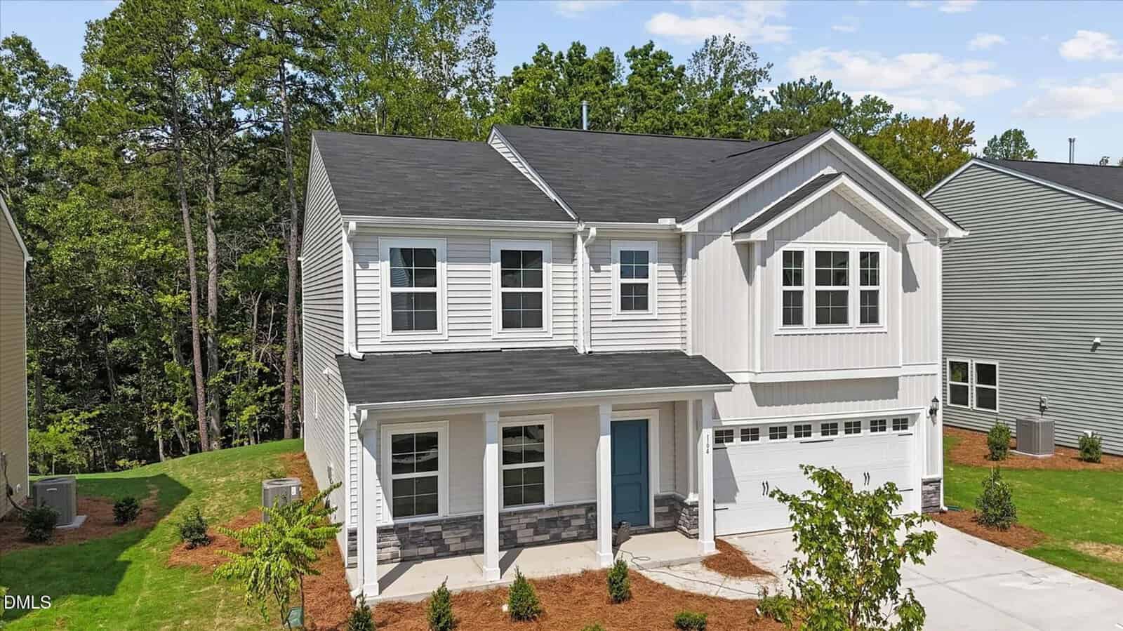 Two-story modern house with white siding, black roof, stone accents, a blue front door, double garage, and a manicured yard, surrounded by trees and neighboring homes on a sunny day.