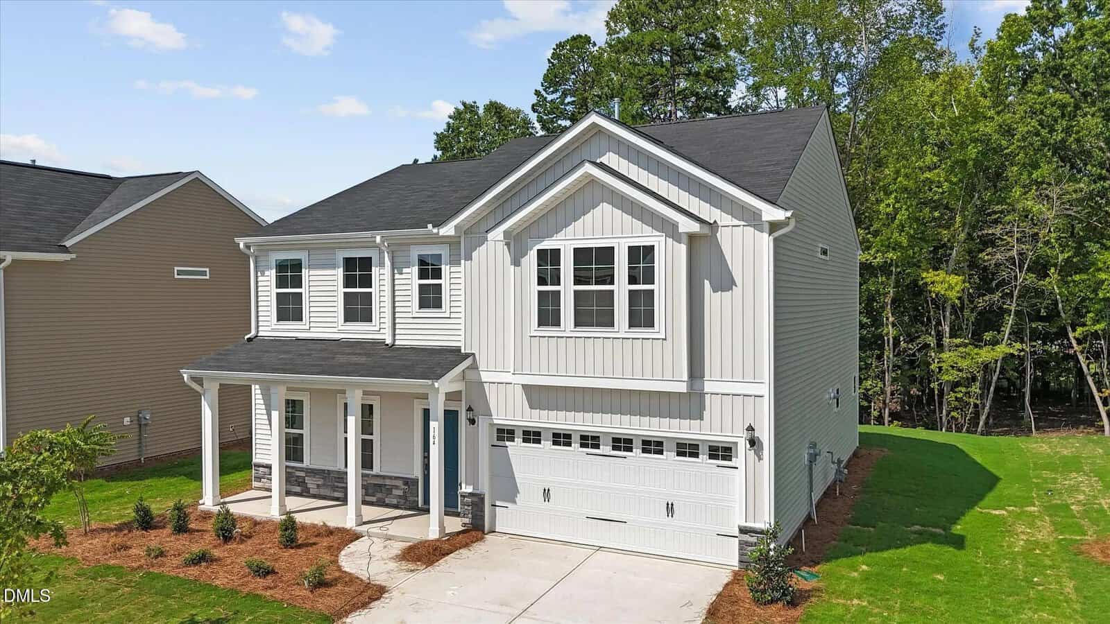 Two-story white house with a covered front porch, double garage, black roof, and neatly landscaped yard, surrounded by green grass and trees under a blue sky.