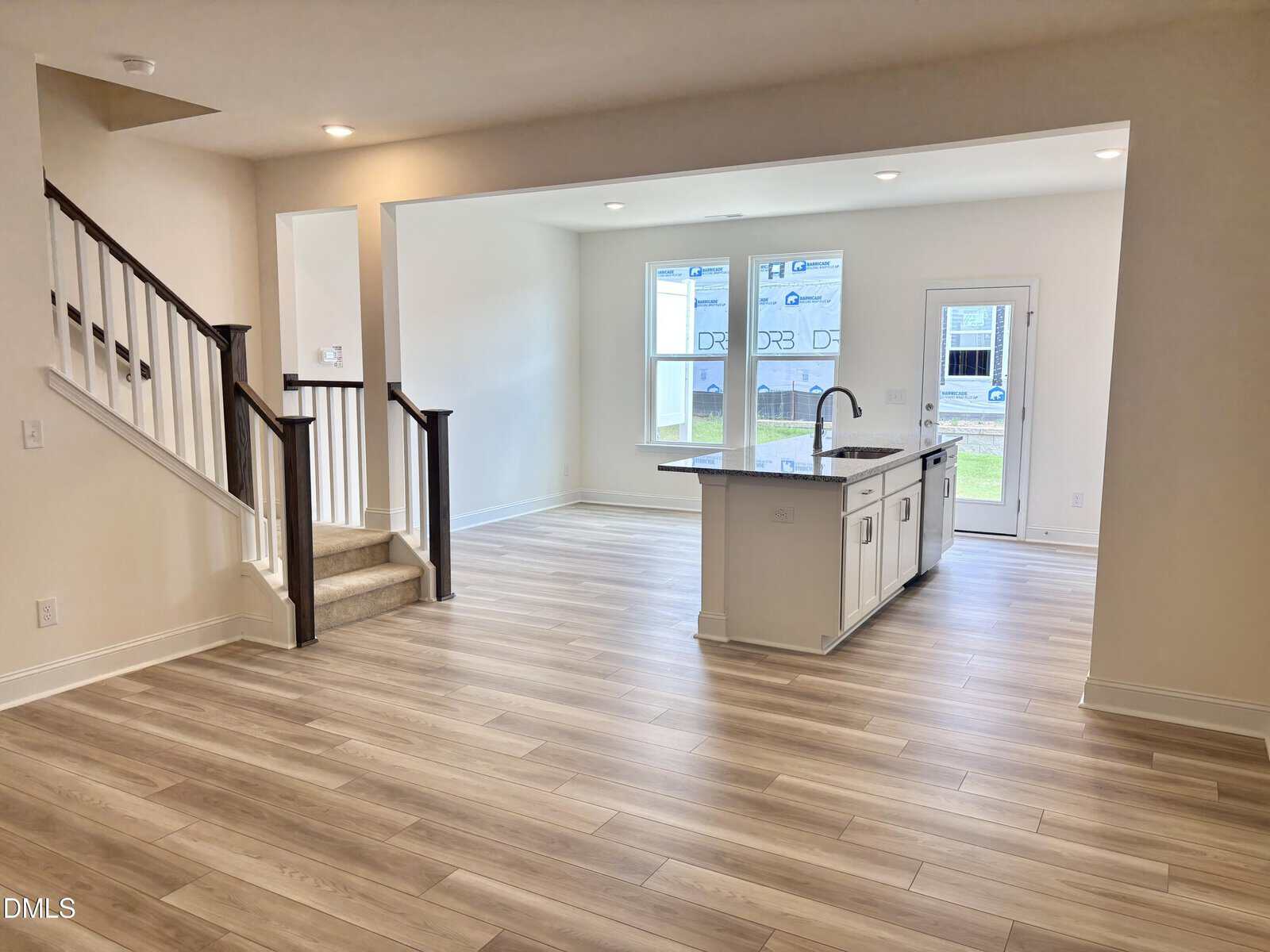 Open-concept living area with light wood floors, an island kitchen, large windows, and a staircase leading upstairs. The space is bright with natural light and has a modern, neutral design.