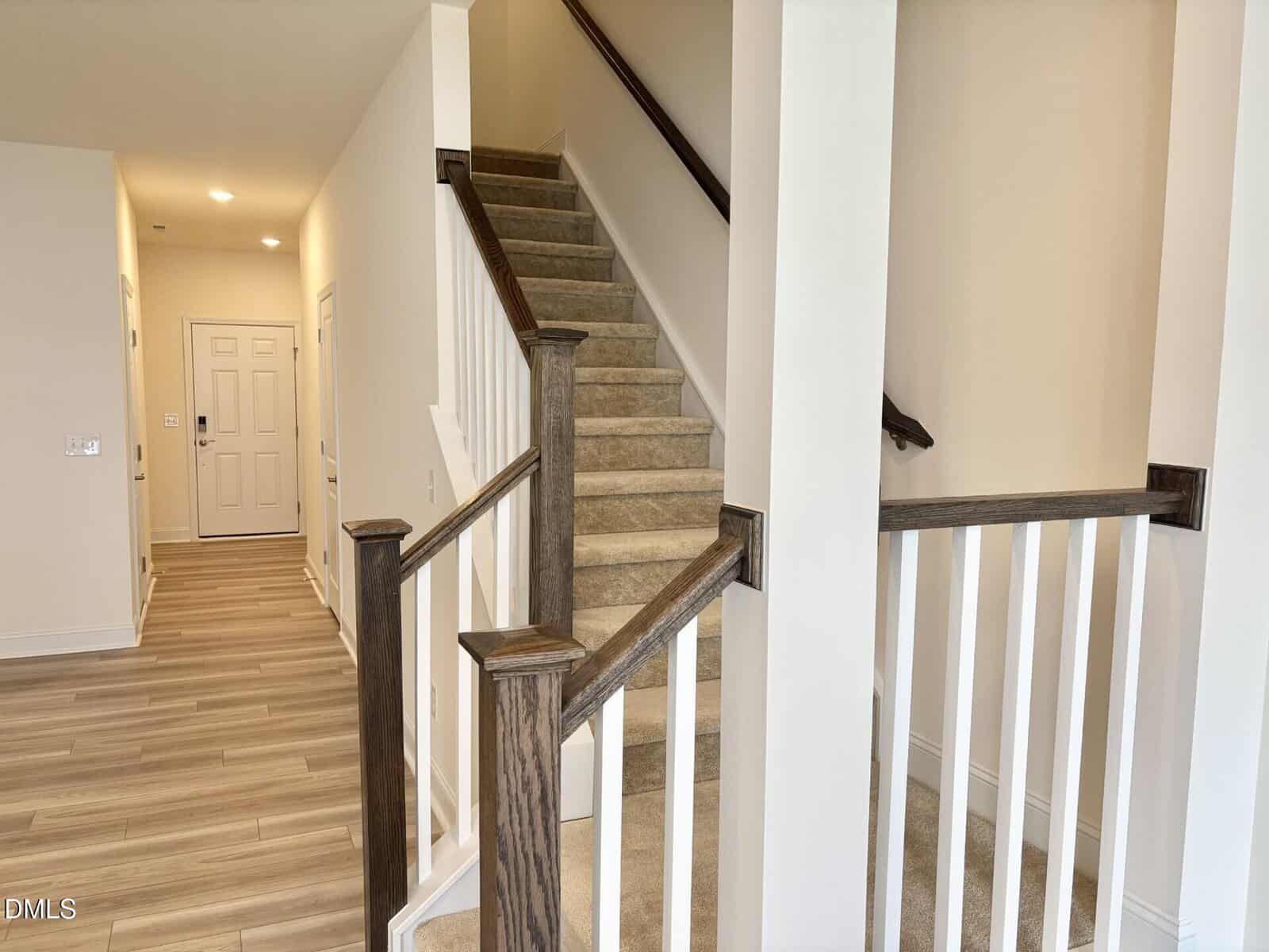 A modern entryway with light wood flooring leads to a carpeted staircase with dark wood railings and white balusters. Walls are painted off-white, and a white door is visible at the end of the hallway.