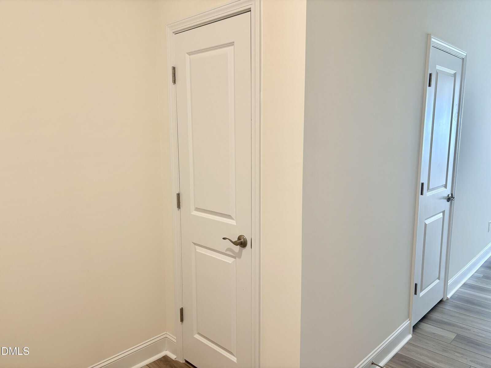 A hallway with light-colored walls and flooring, featuring two closed white doors with silver handles; one door is in the foreground on the left, the other is farther down the hallway on the right.