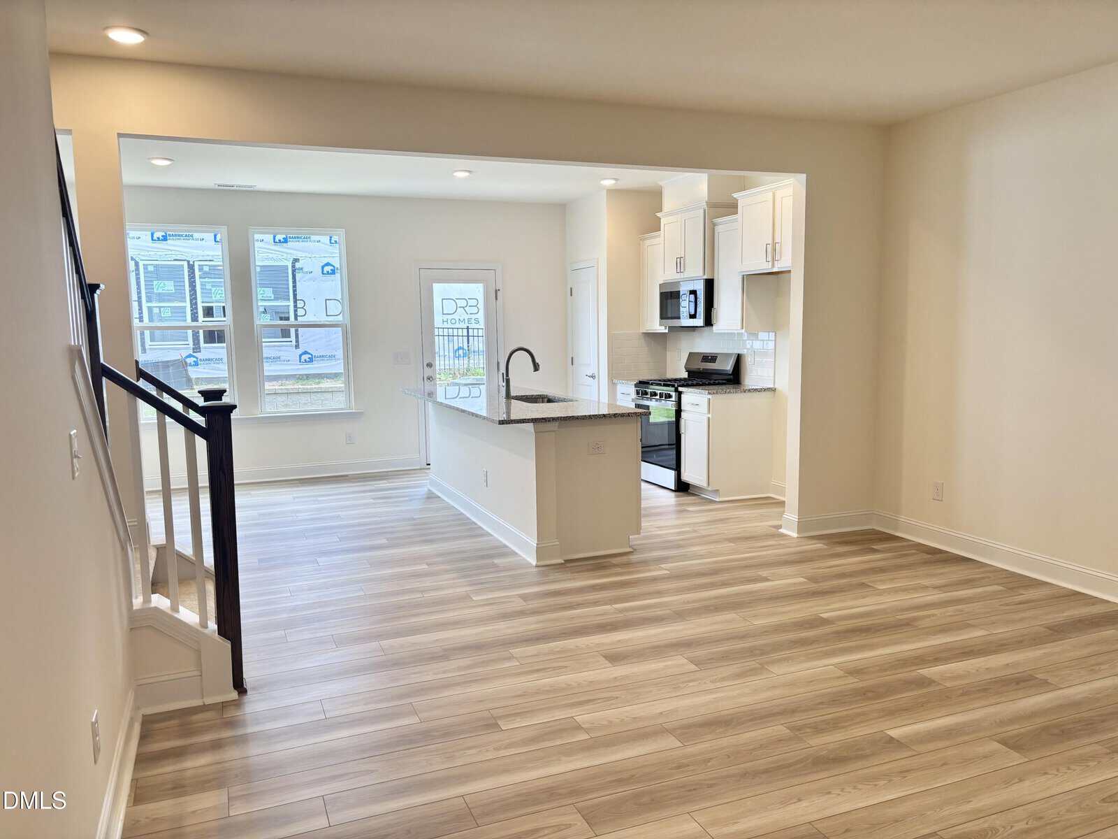 Open-concept kitchen and living area with light wood floors, white cabinets, stainless steel appliances, center island, and stairs on the left. Large windows provide natural light.