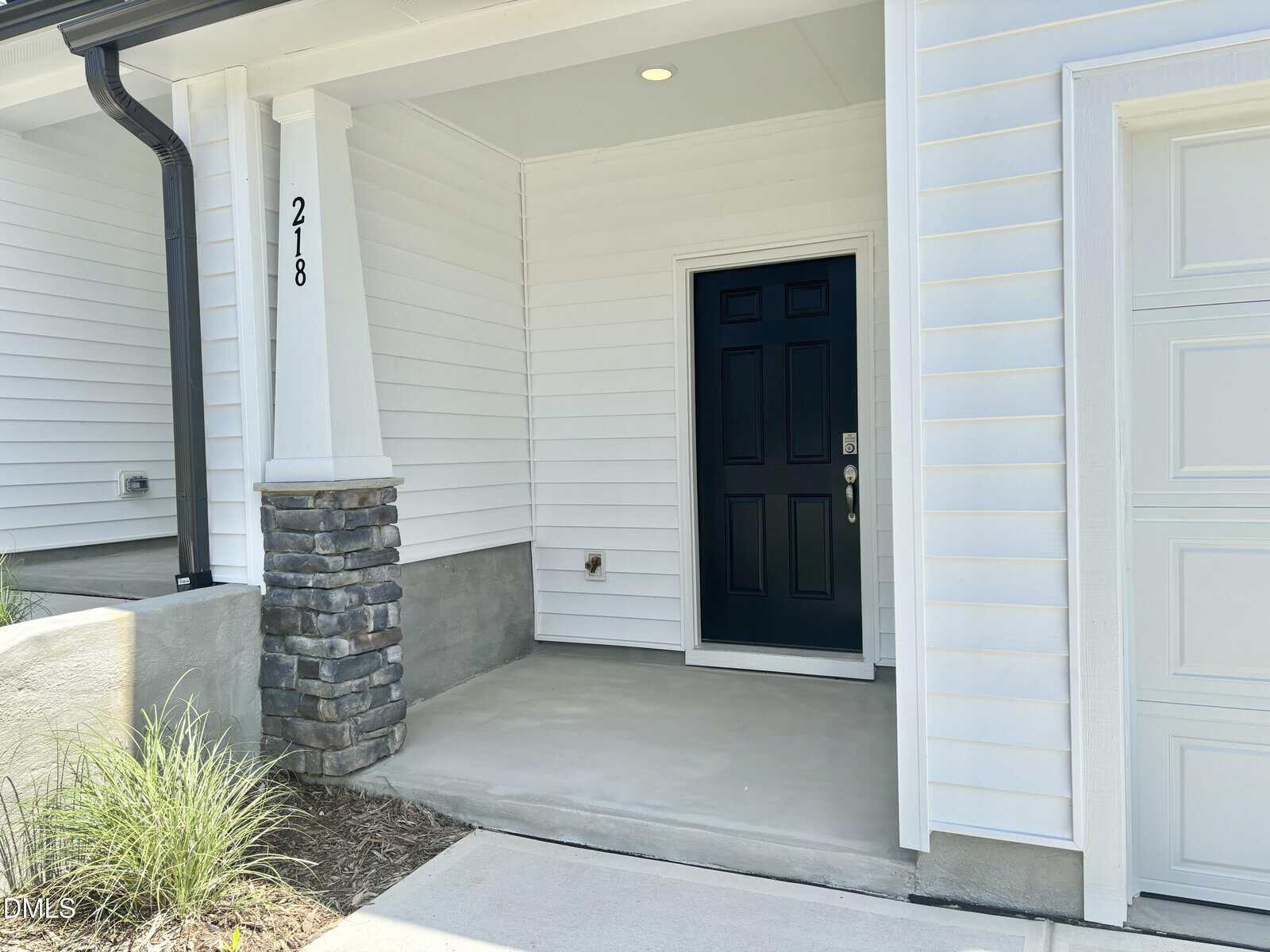 A front porch with white siding, a black door, a single column with stonework at the base, and the house number 218 displayed vertically next to the entrance. A small patch of landscaping is visible on the left.