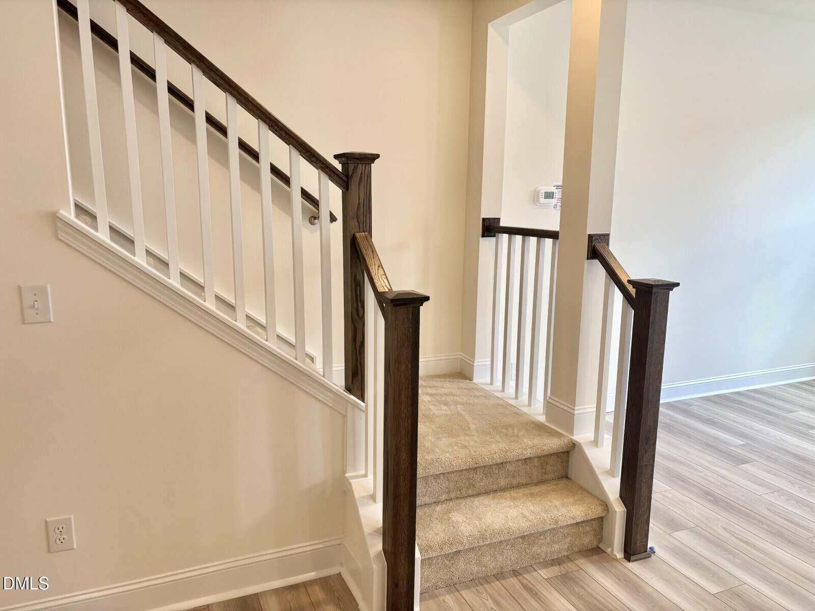 A small set of carpeted stairs with dark wooden handrails and white balusters leads up to a landing. The surrounding walls are light-colored, and the adjacent floor is wood-patterned. A light switch and thermostat are visible.