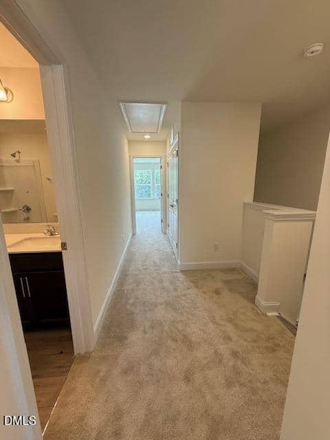 Carpeted hallway with a bathroom on the left, white walls, and a railing on the right. The hallway leads to a well-lit room with large windows at the far end.