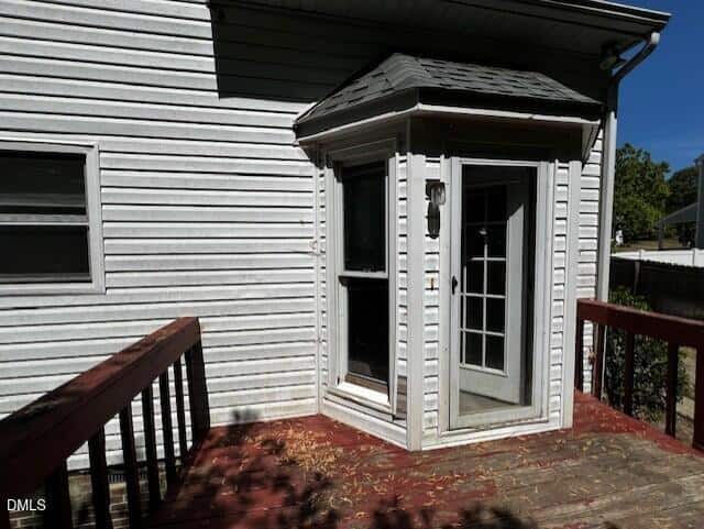 A small, enclosed porch with glass windows and a door is attached to a white house with horizontal siding. The porch opens onto a wooden deck with red railings. Sunlight casts shadows across the deck.