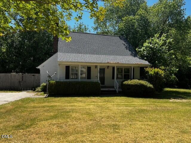 A small white house with a steep roof and front porch sits behind a trimmed lawn and bushes, surrounded by tall green trees under a clear sky.