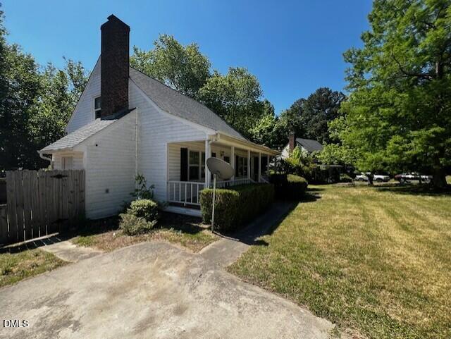 A white house with a brick chimney, front porch, and satellite dish sits next to a grassy yard with trees and a driveway under a clear blue sky.