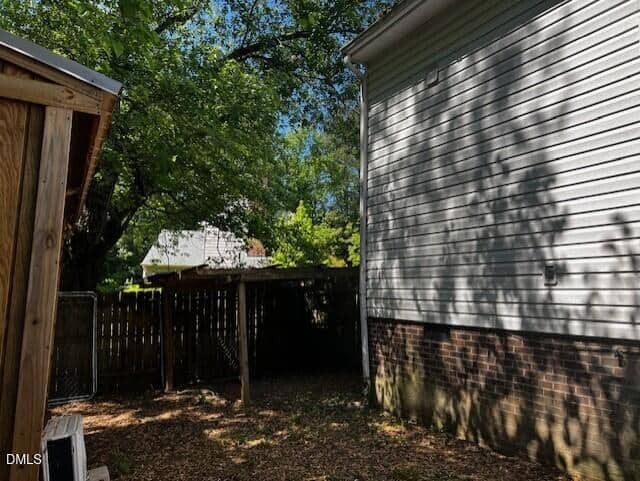 A shaded backyard area with a wooden fence, the side of a house with white siding and brick, a small wooden shed, and trees casting shadows on the ground and wall.