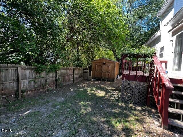A fenced backyard with a small wooden shed, trees providing shade, and red stairs leading up to a house on the right side. The grass is patchy and sunlight filters through the trees.