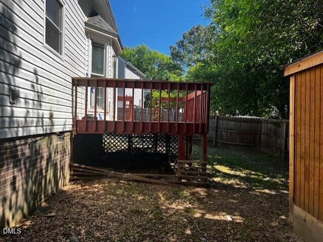A small red wooden deck attached to a gray house, with stairs leading down to a fenced backyard. Trees provide partial shade and the sky is clear and blue.