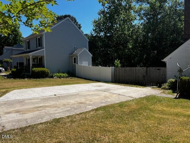 A two-story gray house with white trim is next to a driveway and a white fence, with green grass and trees in the yard on a sunny day.
