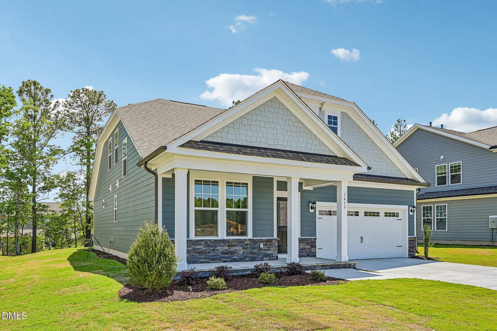 A modern two-story house with blue-gray siding, stone accents, a covered front porch, and a white garage door. The lawn is neatly landscaped with shrubs, and the sky is clear with a few clouds.