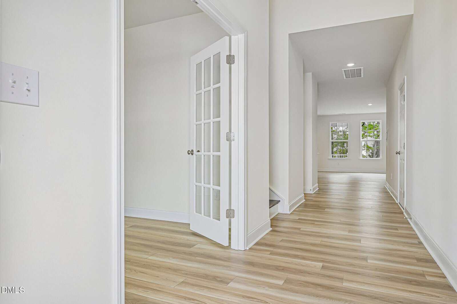 Bright hallway with light wood floors, white walls, a glass-paned French door on the left, and an open view to a living area with large windows in the background.