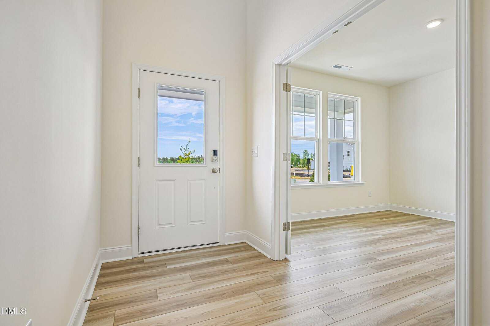 Bright entryway with light wood floors, white walls, a white front door with window, and an adjacent room with large windows letting in natural light. Outdoor scenery is visible through the windows.