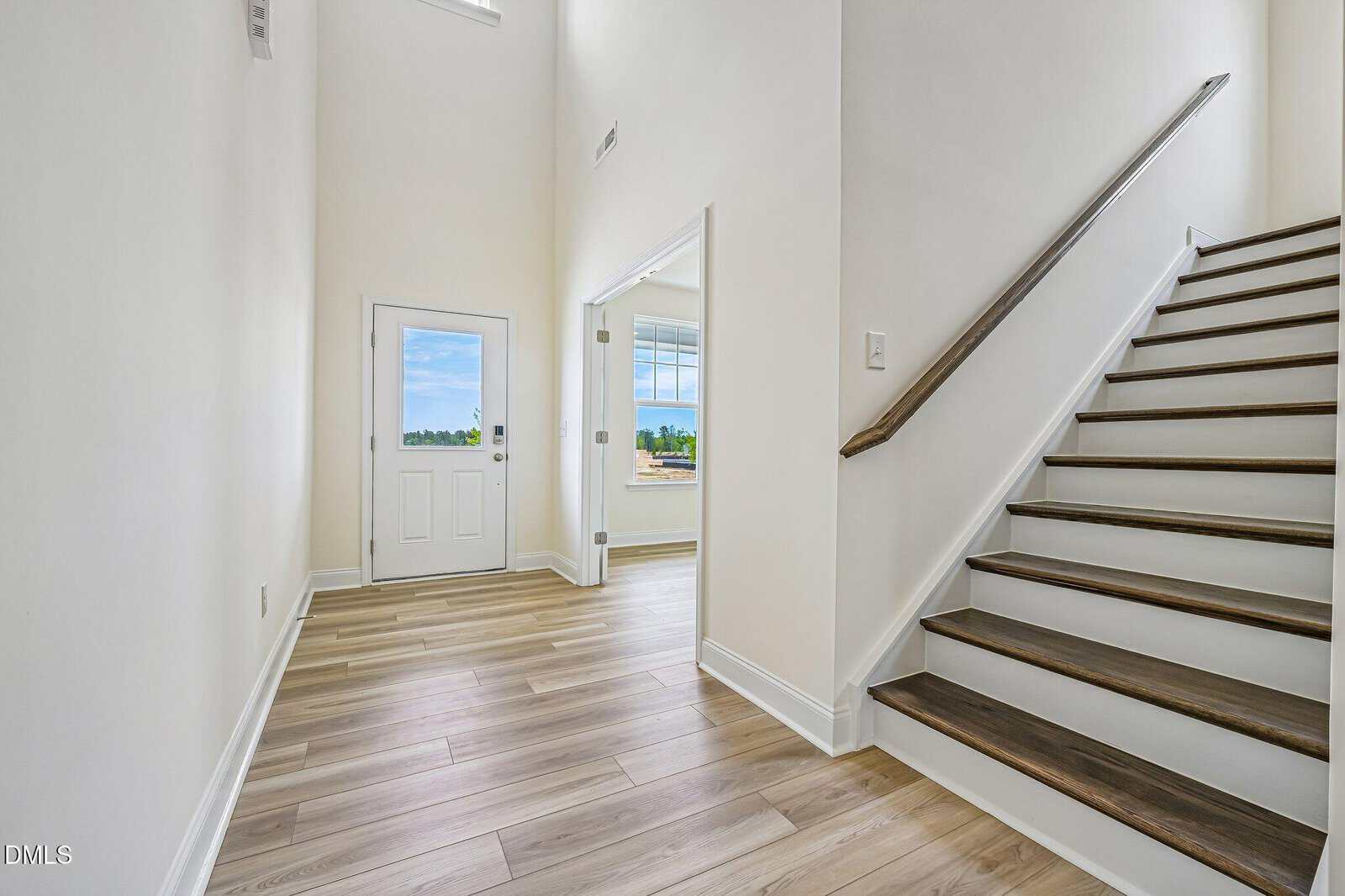 Bright entryway with light wood flooring, white walls, a staircase with dark wood steps on the right, and a white front door. Sunlight streams in from windows in the adjoining room and above the door.