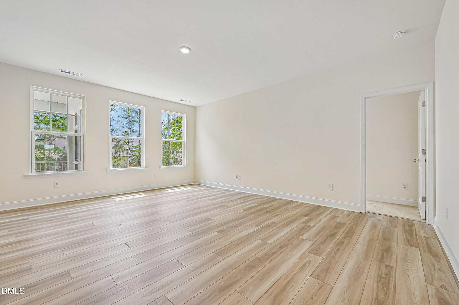 Bright, empty room with light wood flooring, white walls, three large windows letting in natural light, and a single doorway leading to an adjacent room.