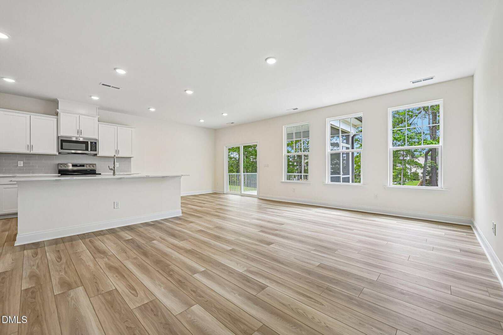 Spacious, modern open-concept living area with light wood floors, white walls, large windows, and a kitchen with white cabinets and stainless steel appliances.