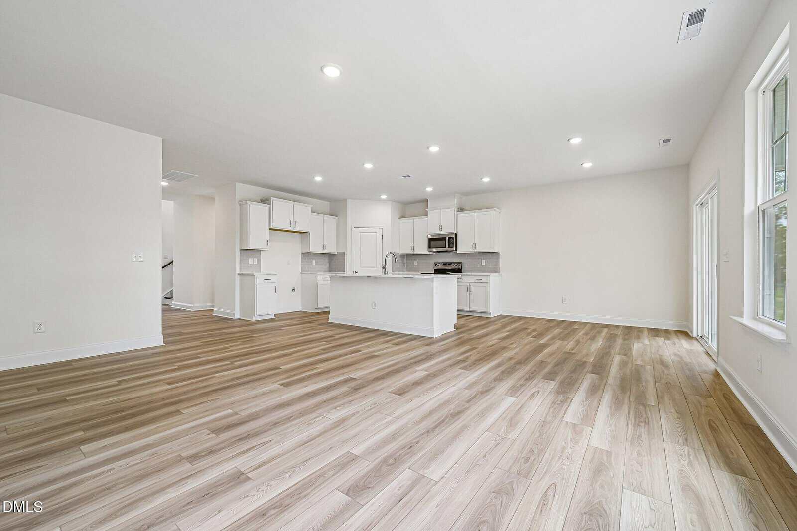 Spacious modern kitchen with white cabinets, island, and stainless steel appliances open to a large living area with light wood flooring and ample natural light from large windows.