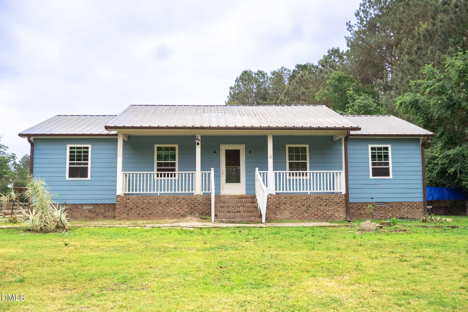 A single-story blue house with white trim and a metal roof, featuring a covered front porch with white railings and brick steps, set in a grassy yard with trees in the background.