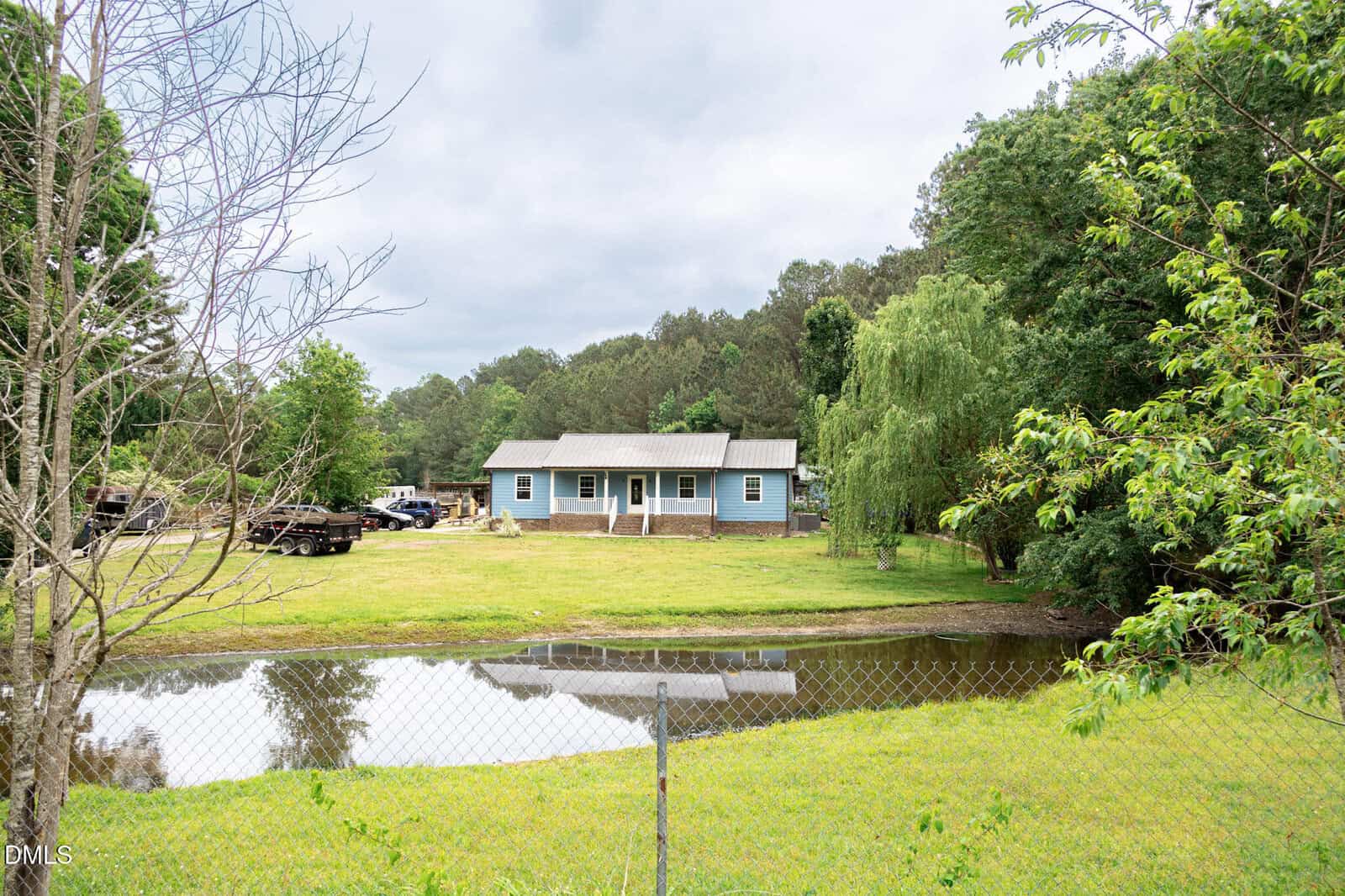 A single-story gray house with a front porch sits in a grassy yard surrounded by trees, with a small pond and a chain-link fence in the foreground. Several vehicles are parked to the left of the house.