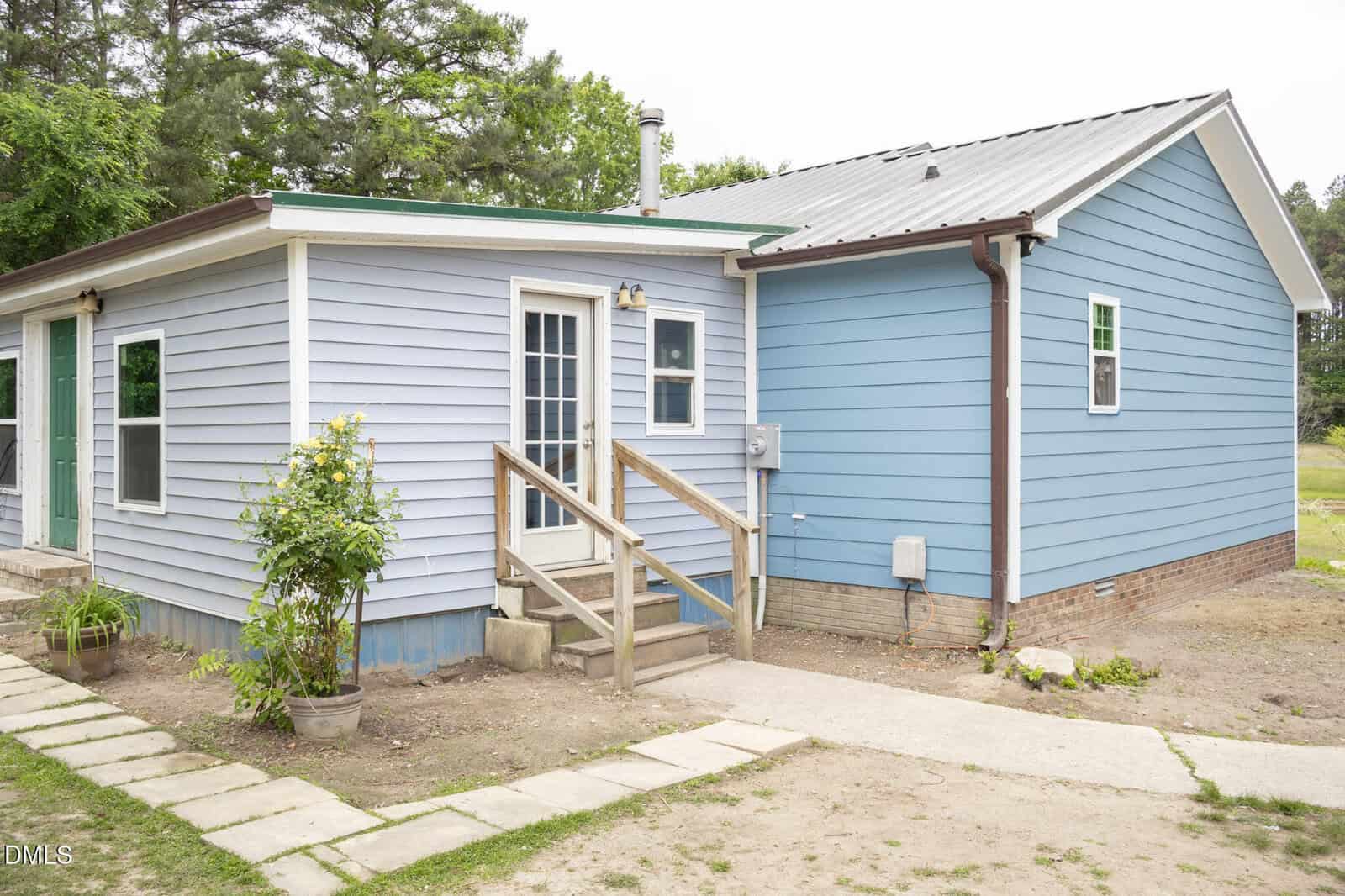 Single-story house with light blue and darker blue siding, a small wooden staircase leading to a white door, potted plants by the entrance, brick foundation, and a grassy yard with trees in the background.