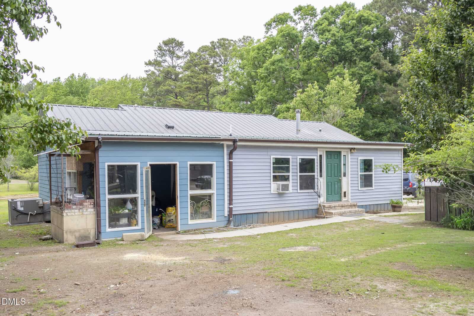 A single-story blue house with a green front door, multiple windows, and double glass doors opening to a yard with patchy grass and trees in the background.
