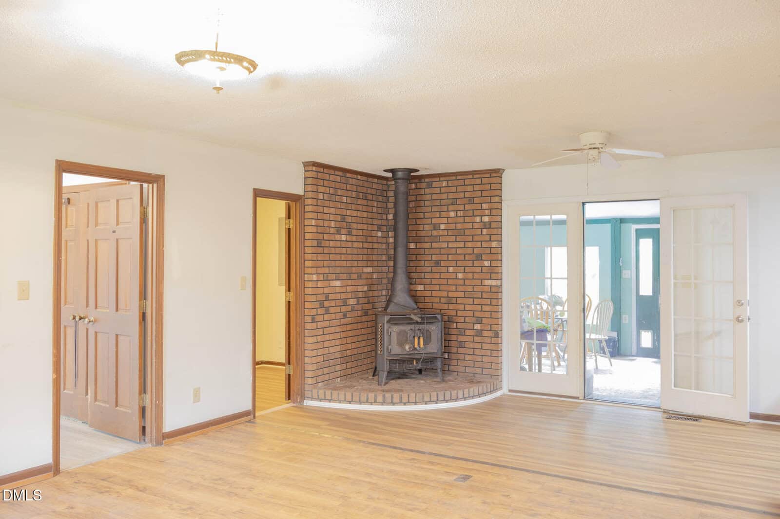 Bright living room with wood flooring, a vintage wood-burning stove set in a brick corner, a ceiling fan, and double glass doors opening to a sunroom with a dining table and chairs. Multiple wooden doors lead to other rooms.