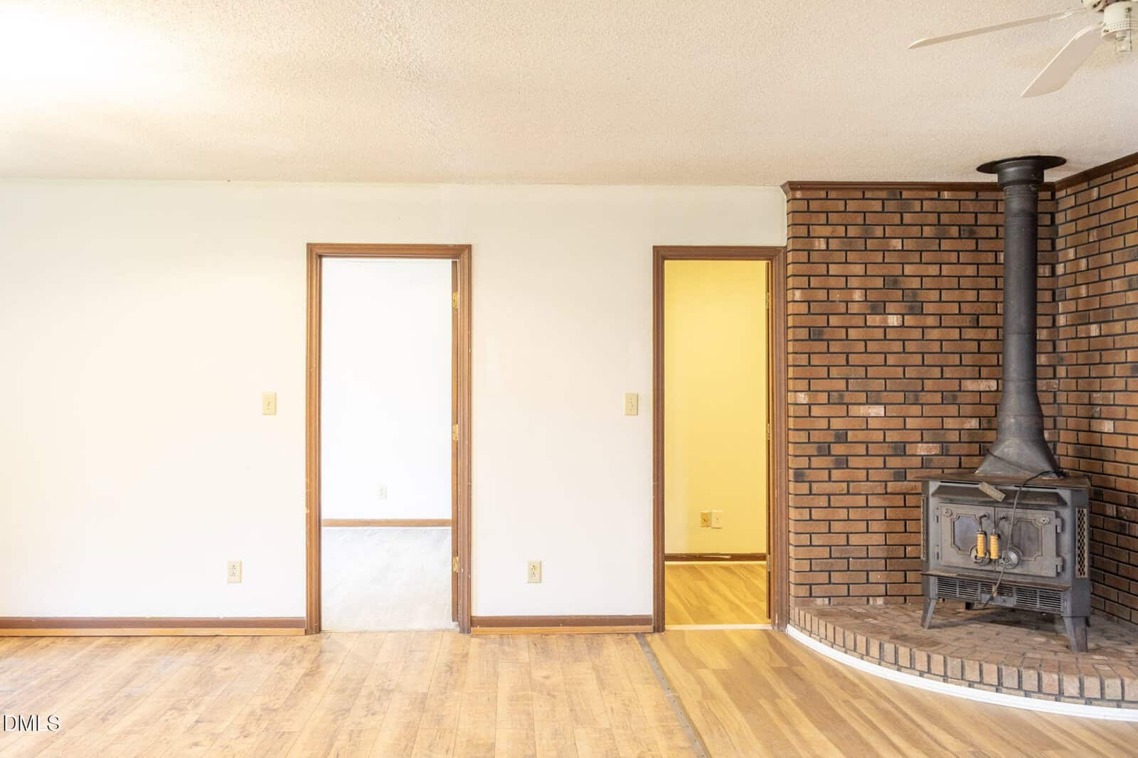 A room with light wood floors, two doorways in a white wall, and a brick corner with a black wood stove and vent pipe on a raised brick hearth. Ceiling fan partially visible.