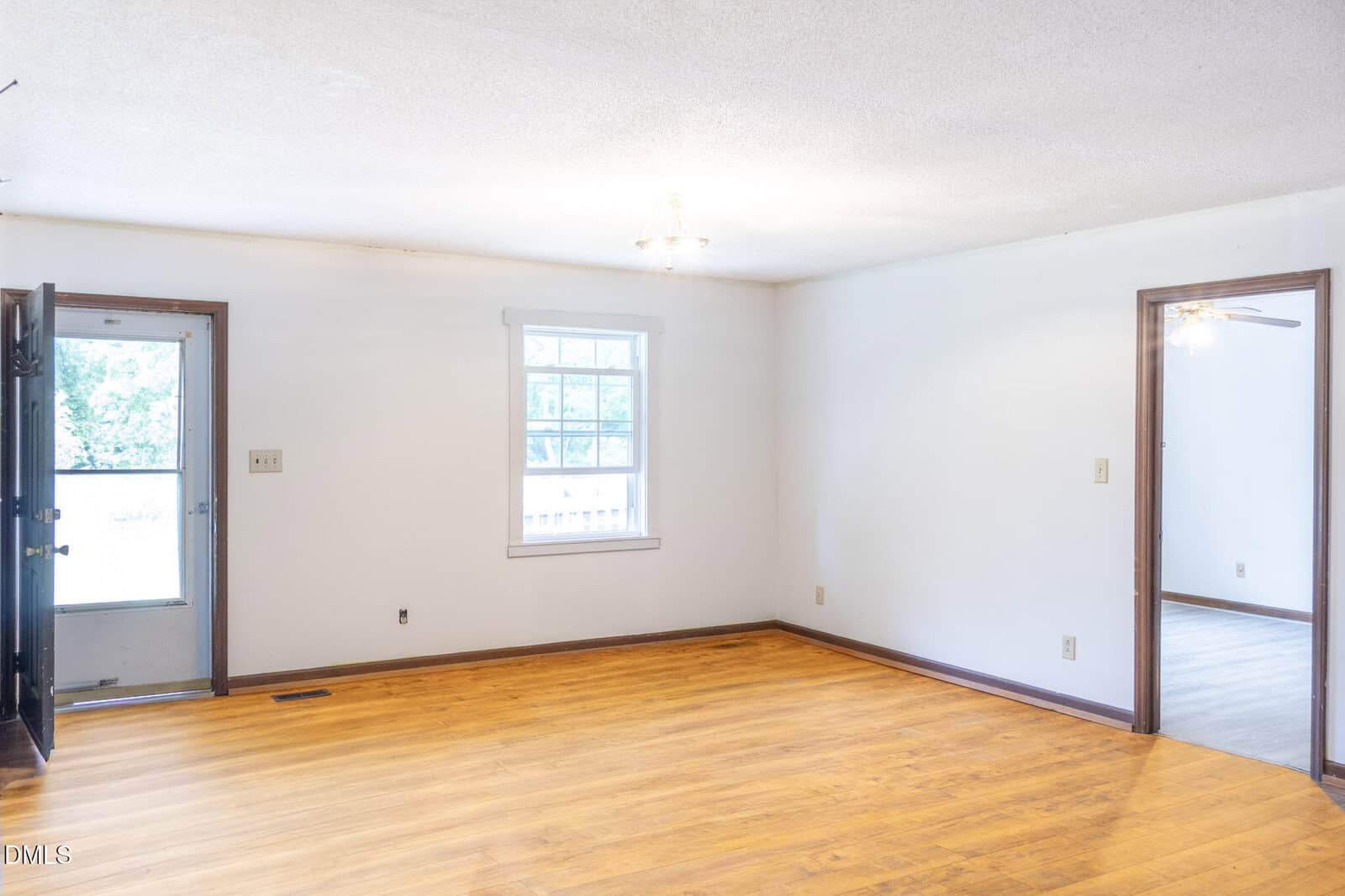 Empty living room with white walls, a window, wooden floor, dark trim, a ceiling light, open front door, and an open doorway leading to another room with a ceiling fan. Natural light fills the space.