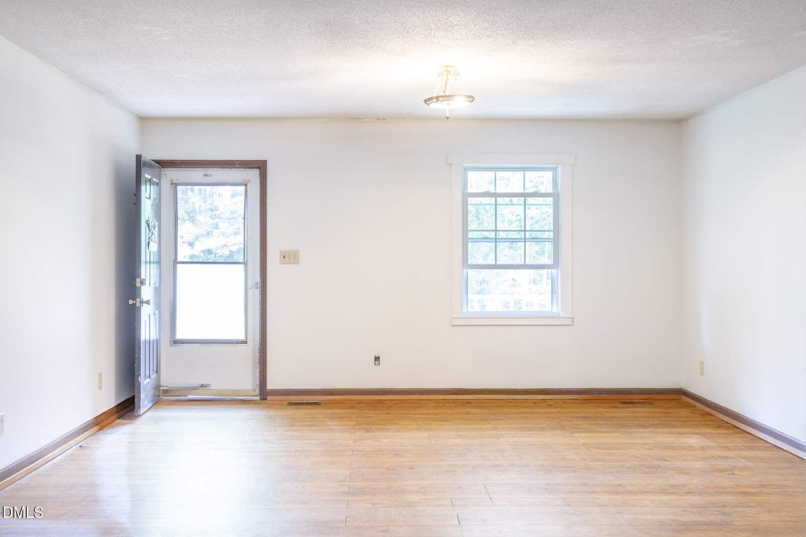Empty room with light-colored walls, wood floor, a window on the right wall, and an open door on the left wall. Ceiling light fixture in the center. Natural light is coming through the window and door.