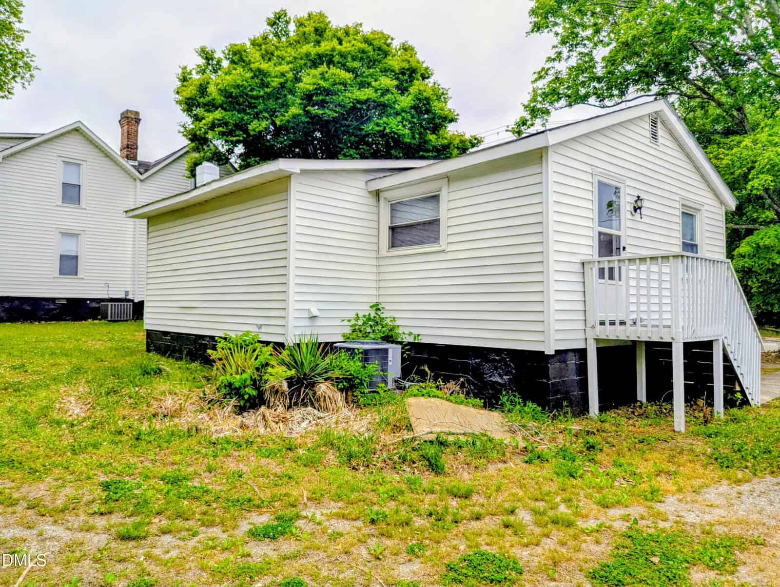 A small white house with black foundation, white siding, a single window, and a short staircase leading to a side door. There are some green bushes and trees surrounding the yard.