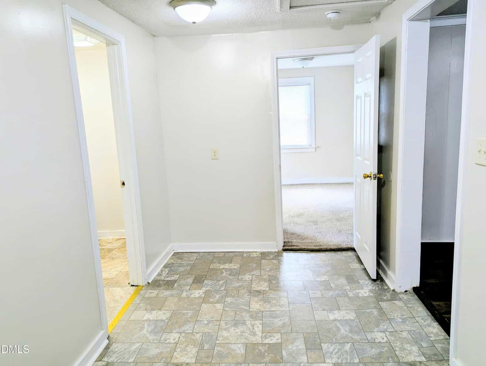 A small hallway with tile flooring connects to three rooms. One room has carpet, one has tile, and the third room is only partially visible. All walls are white with a ceiling light fixture.
