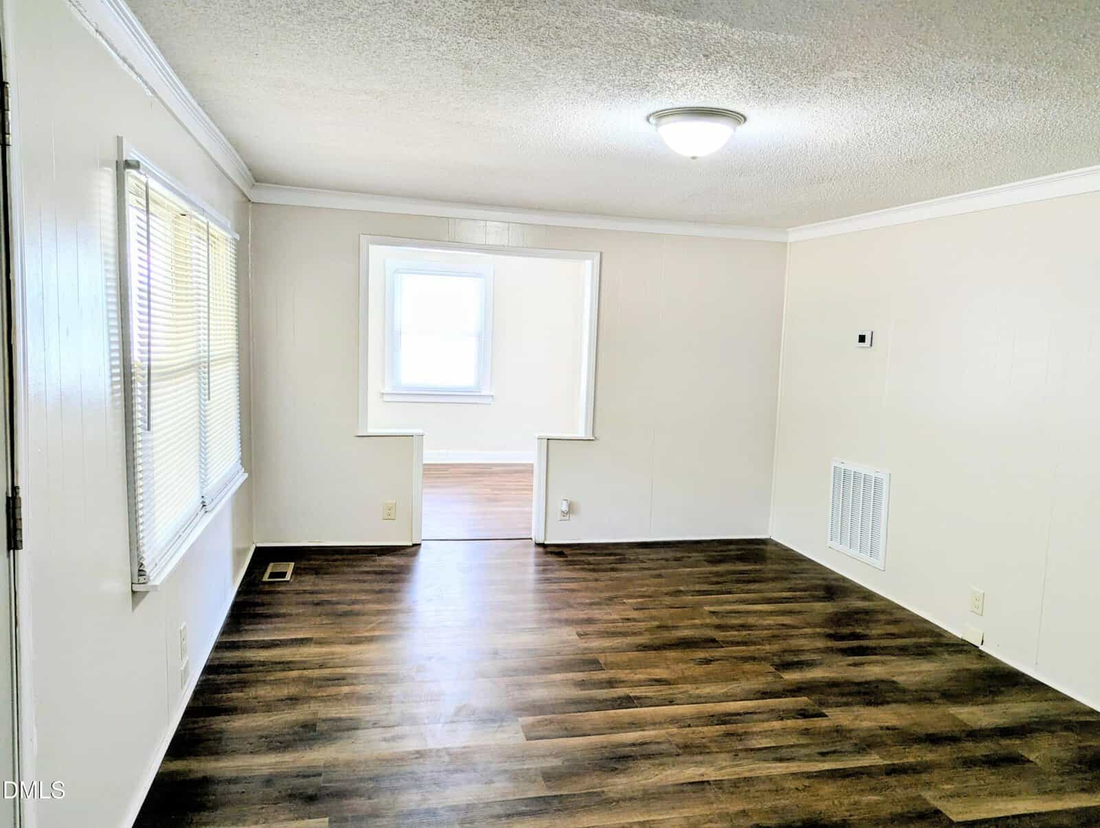 Bright room with white walls, dark wood flooring, a ceiling light, and two large windows. An open doorway leads to another well-lit room with similar flooring and a large window.