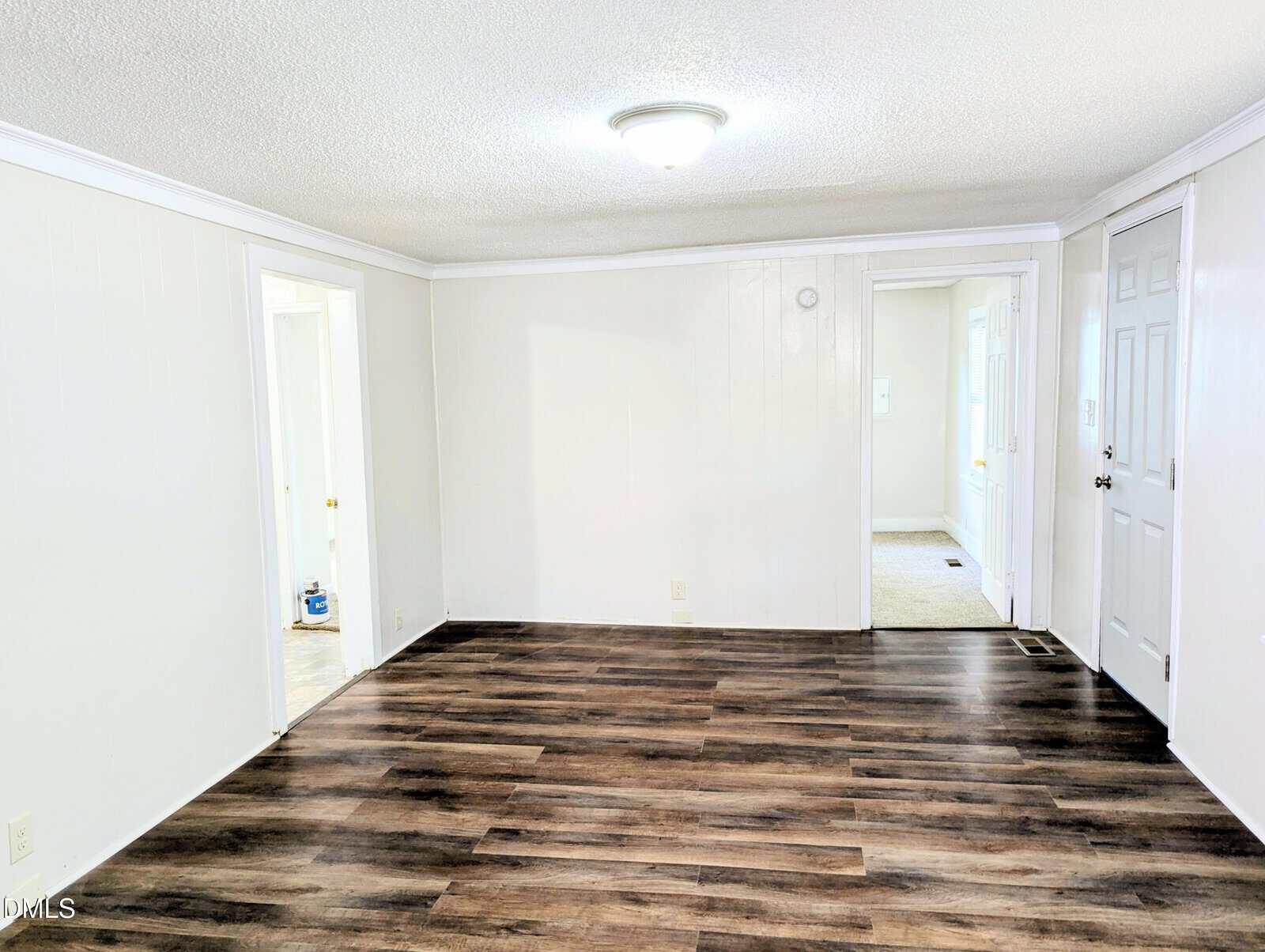 Bright, empty room with white walls, dark wood flooring, a ceiling light fixture, and multiple doorways leading to other rooms. One door is closed and painted white. The space appears clean and freshly renovated.