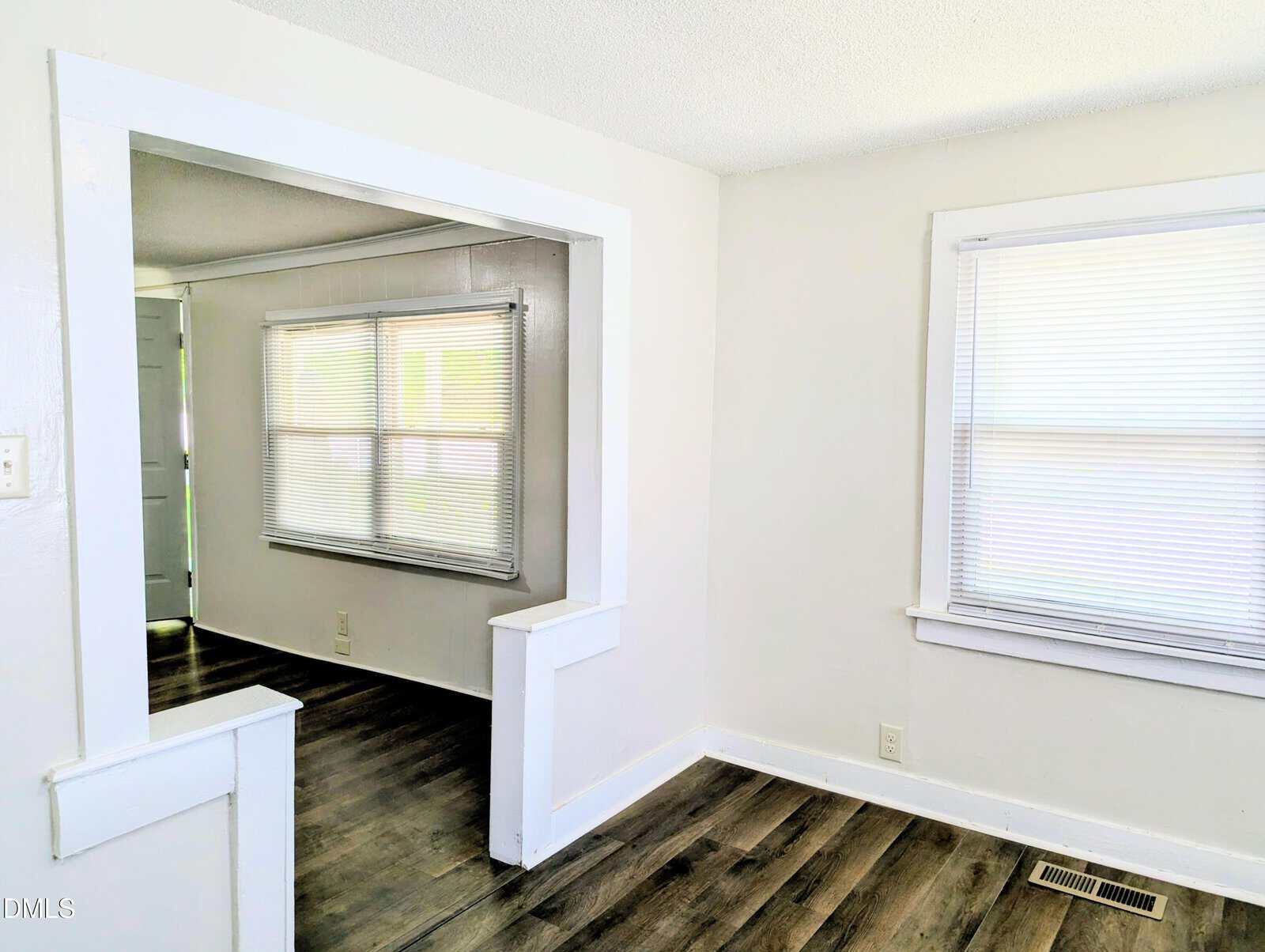A bright, empty room with white walls and blinds covering the windows. The room has dark wood flooring and an open doorway leading to another similar room, also with windows and blinds.