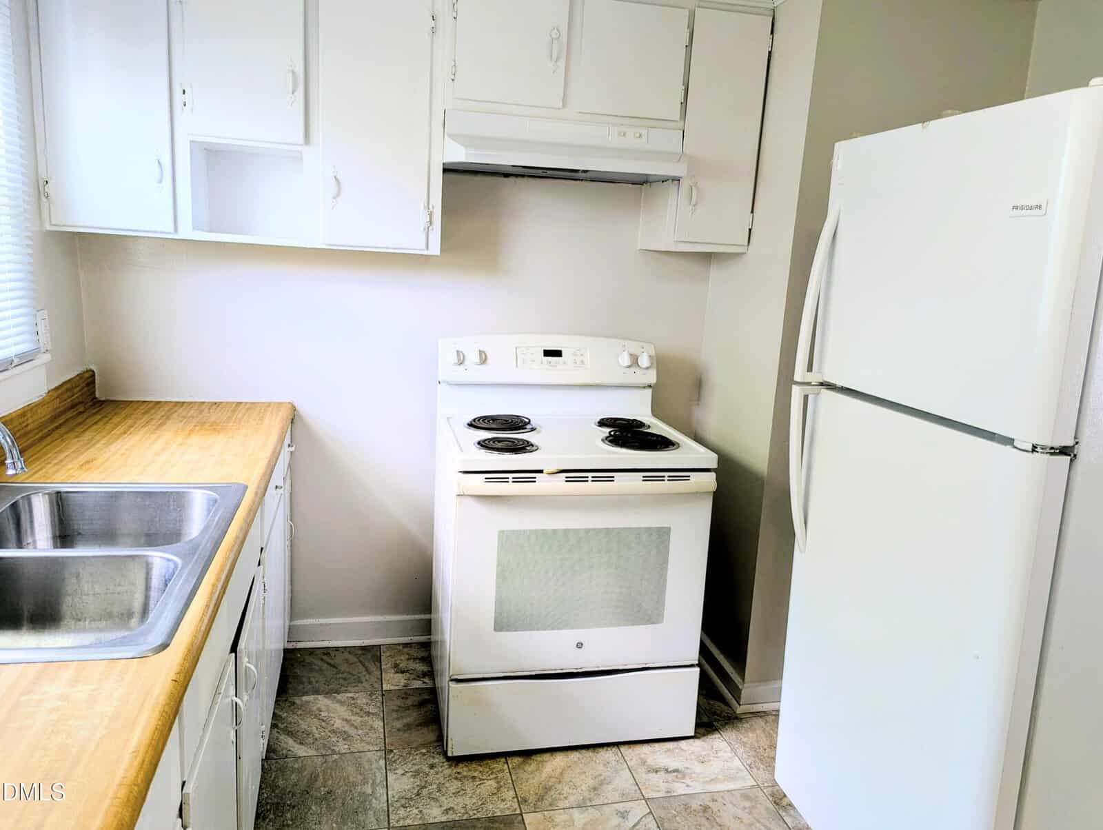 Small kitchen with white cabinets, electric stove, white refrigerator, double sink with metal faucet, wood-look countertops, and tiled floor. The walls are light-colored, and there is a window above the sink.