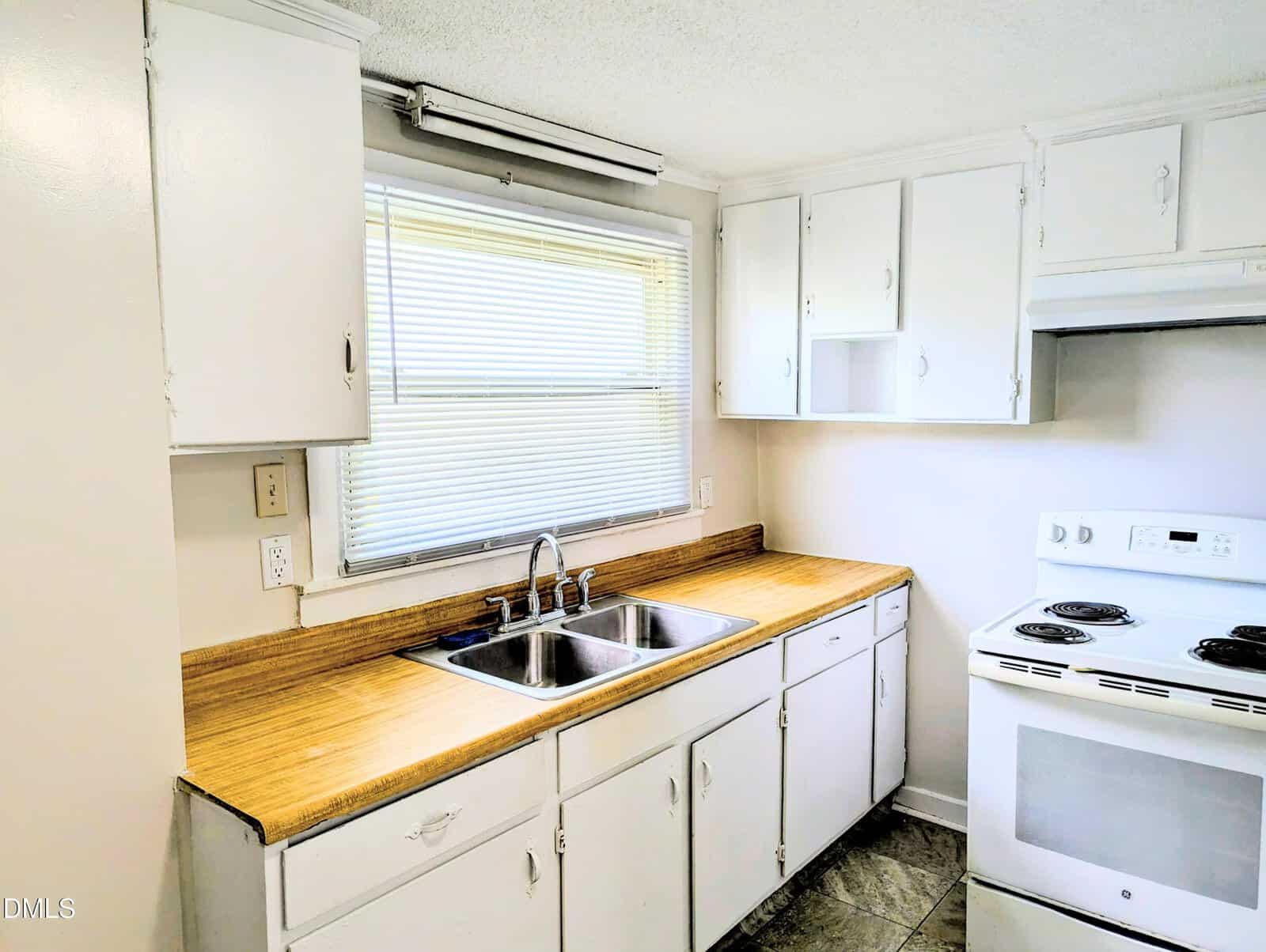 A small kitchen with white cabinets, a double stainless steel sink under a window with blinds, wood countertops, and a white electric stove with a range hood on the right side.
