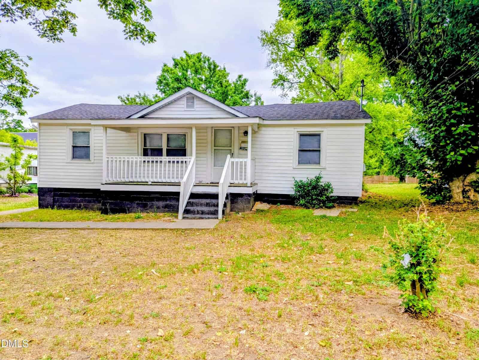 Charming single-story white house at 908 S Main Street, Louisburg, with a small front porch, black foundation, and gray roof. Surrounded by grass and trees, it features minimal landscaping plus a ramp and stairs leading to the porch.