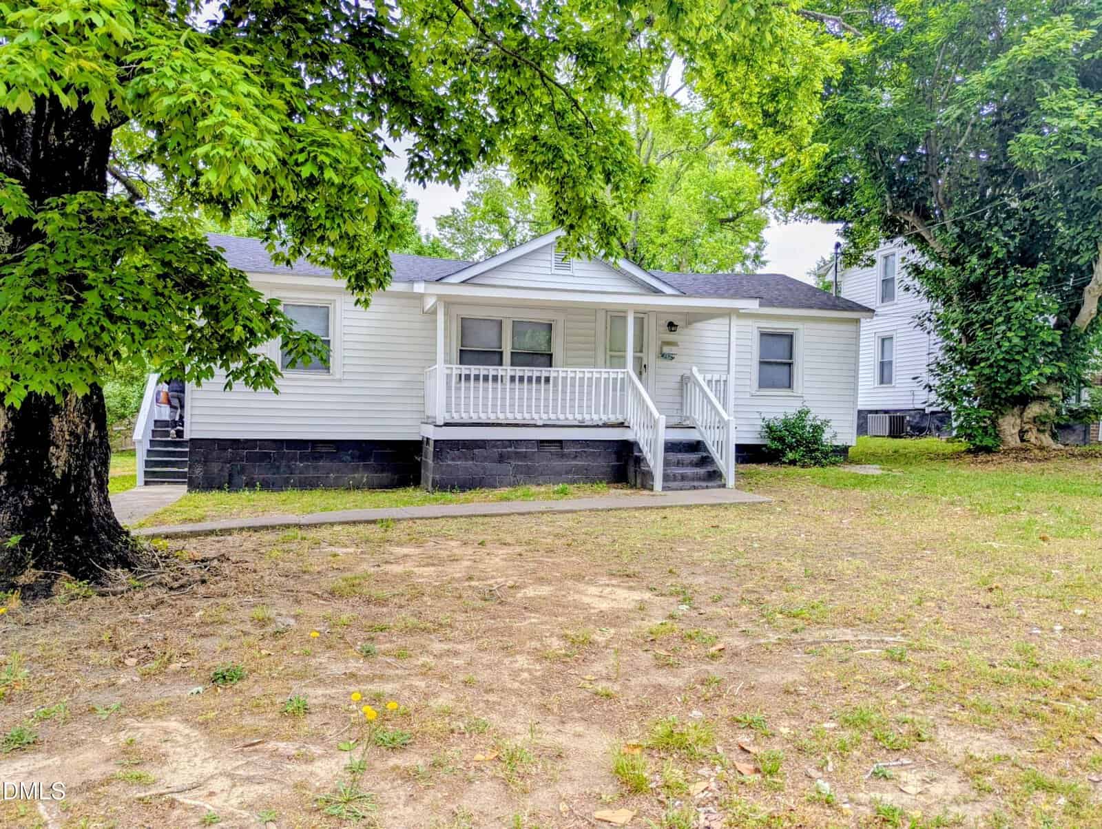 A small white single-story house with a covered front porch and steps, surrounded by trees and a patchy grass yard. The house has black trim along the foundation and a concrete walkway leading to the porch.