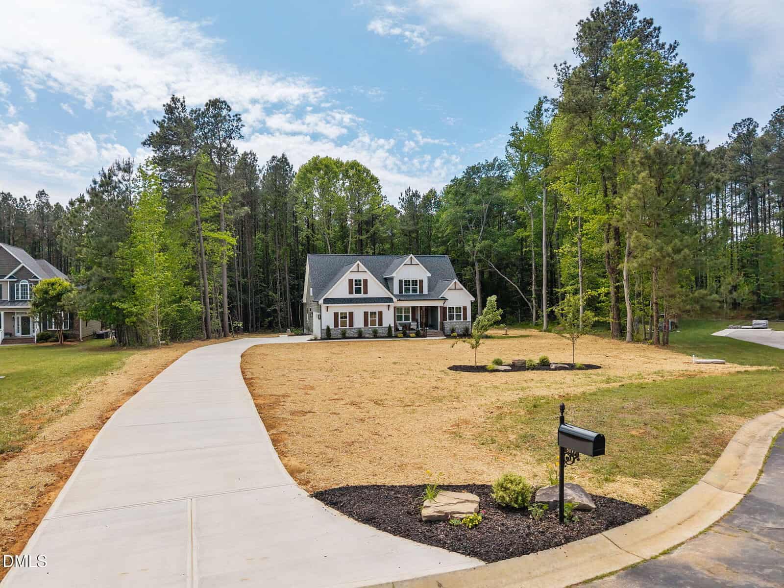 A two-story suburban house with a gray roof and white siding sits at the end of a long, curved driveway, surrounded by a large lawn with sparse grass and bordered by trees. A black mailbox is near the road.