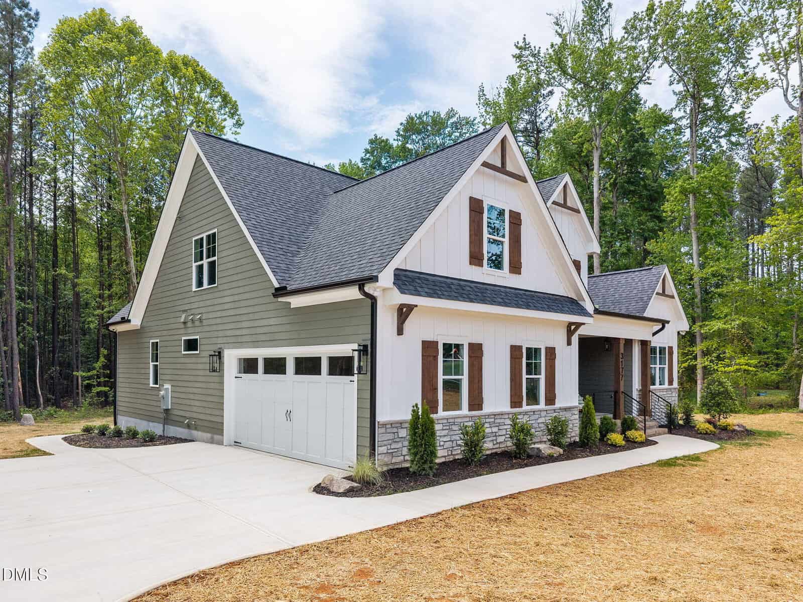 Modern suburban house with light gray siding, white trim, stone accents, and a gabled roof. A double garage door faces a wide driveway, and simple landscaping lines the front. Surrounded by trees and grass.