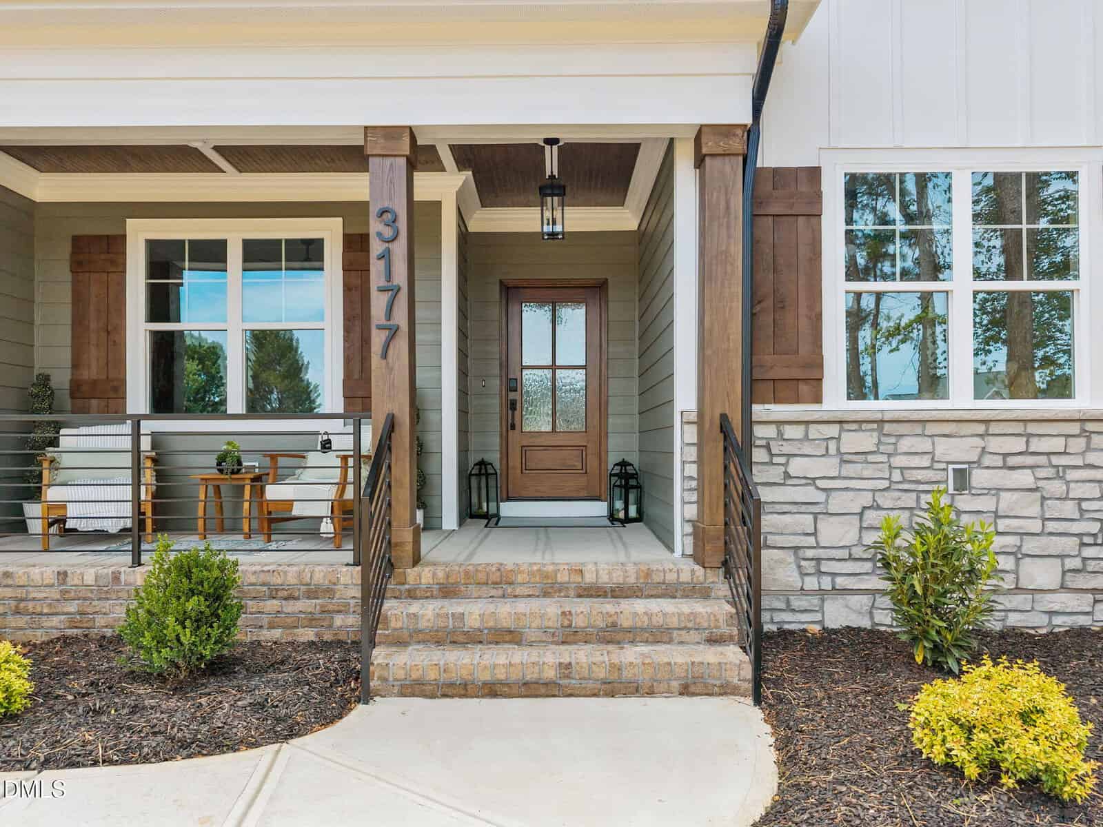 Front entrance of a modern house with a wooden front door, large windows, porch seating area, house number 3177 on the pillar, brick steps, metal railing, and landscaped garden beds.