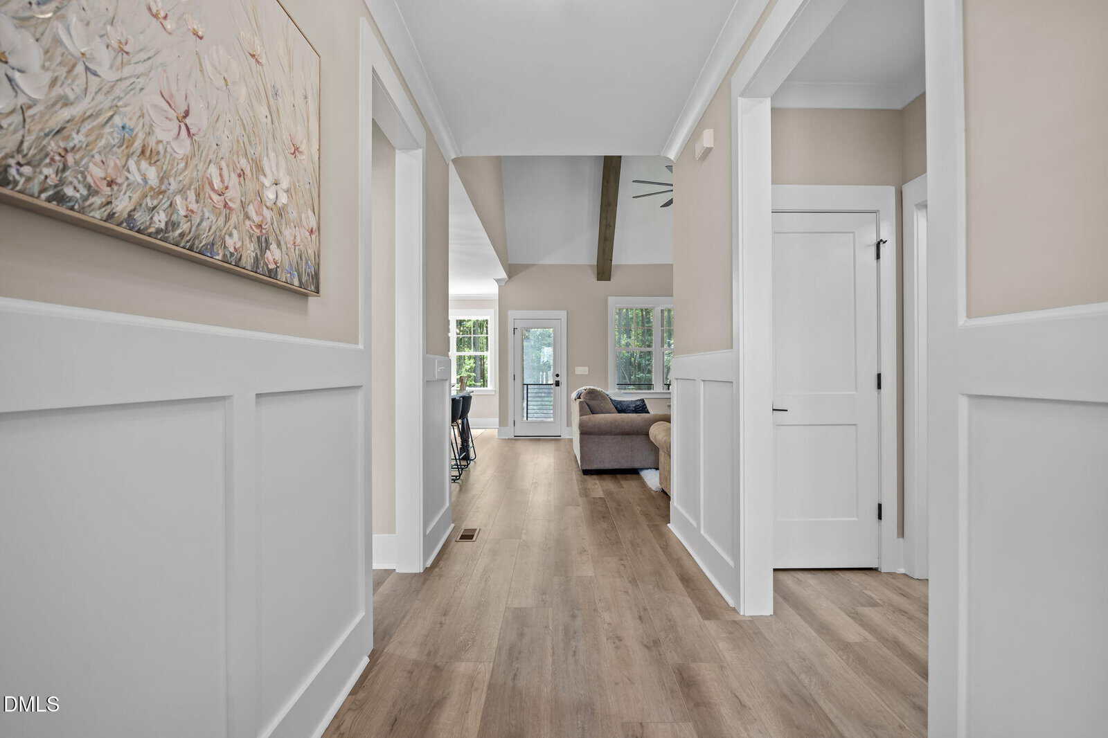 Bright hallway with light wood flooring, white paneled walls, and artwork on the left. The hallway opens to a living area with a sofa, visible windows, and a glass door leading outside.