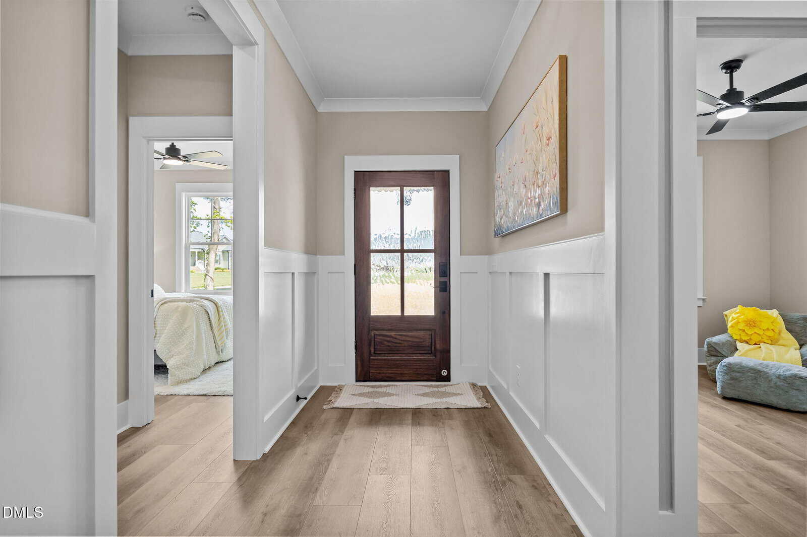 Bright hallway with wooden floors, a glass front door, white trim, and beige walls. Rooms on both sides are visible, one with a bed and one with a couch and yellow pillow. A large painting hangs on the right wall.