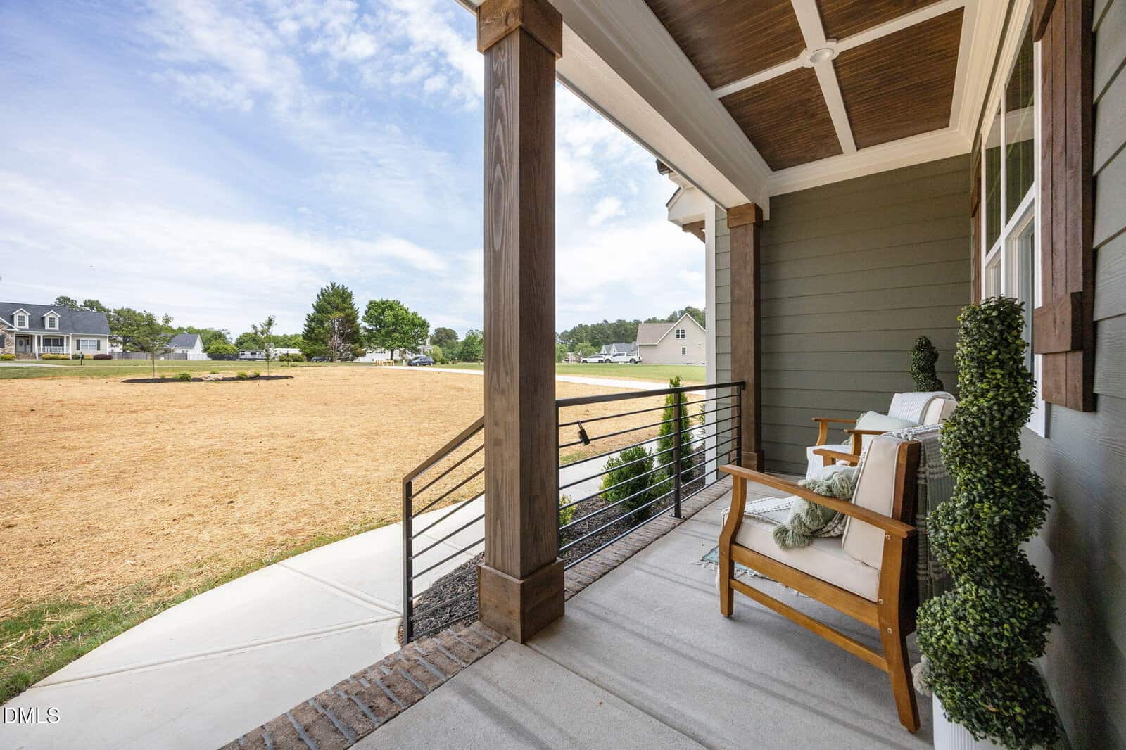 A front porch with two cushioned chairs, decorative plants, and a wooden railing overlooks a lawn under a partly cloudy sky in a suburban neighborhood.
